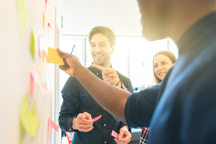 Person pulling a sticky note off a board with another person smiling beside him